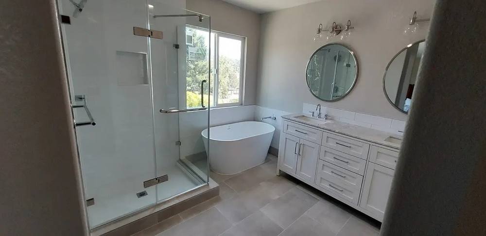 Wide view of a remodelled bathroom: frameless glass shower, freestanding soaking tub, double vanity with round mirrors.