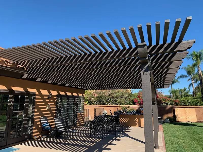 Large timber pergola and patio cover over a paved terrace, sun-louvre shadows, garden and palms behind.